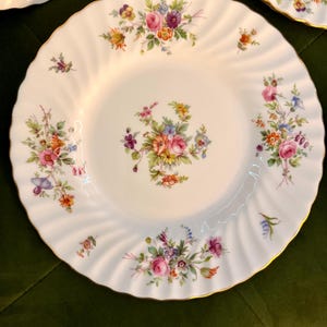 May include: A white porcelain plate with a scalloped, fluted edge and a gold-trimmed rim. The center features a colorful floral bouquet, with additional smaller floral clusters arranged around the border in shades of pink, purple, orange, and green.