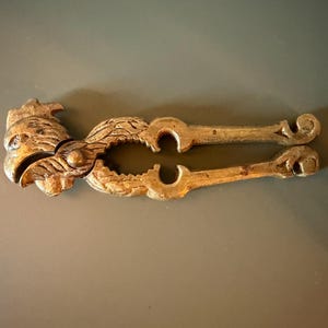 May include: A metallic nutcracker featuring a detailed rooster head design at the hinge. The tool has two long, textured handles with curved ends and a central circular opening for cracking nuts, finished in a weathered, antique gold-toned metal.