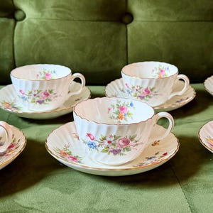 May include: A set of five white porcelain teacups and matching saucers, each featuring a delicate ribbed texture and a colorful floral pattern of pink, purple, and blue flowers with green leaves, finished with a thin gold trim along the edges.