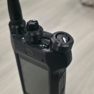 May include: A black two-way radio featuring a textured antenna, a central volume knob, and a secondary dial on the right side. The device includes a small screen, a Motorola logo, and a switch labelled with the letters B and C near the top controls.
