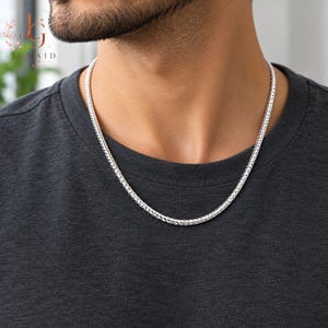 Puede incluir: Un collar de cadena de tono plateado con un patrón de eslabones redondeados y tejidos apretados, tipo trigo o cola de zorro. La cadena metálica descansa sobre una camiseta gris oscuro, mostrando una textura pulida y uniforme en toda su longitud.