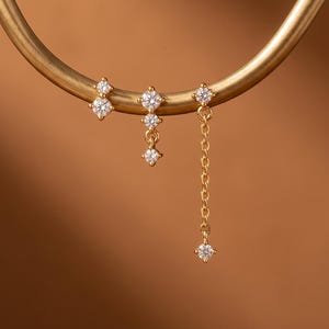 May include: Three gold-tone earrings featuring clear, round-cut stones. The first earring has two stacked stones, the second has three stones in a vertical drop, and the third features a single stone attached to a delicate gold chain with a stone end.