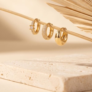 May include: Three gold hoop earrings displayed on a thin wooden stick. The first earring features a central stone with small accents, the second has a pavé-style surface with tiny stones, and the third is a smooth, polished gold hoop earring.