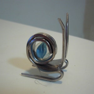 May include: A silver snail sculpture made from a bent fork and spoon with a blue marble for the shell.