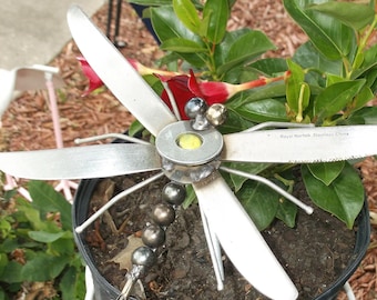 Pode incluir: Uma escultura artesanal de libélula de metal com asas de lâmina de faca, corpo de contas e centro amarelo, colocada em um vaso preto com folhagem verde. O vaso está em um suporte de planta de metal branco. O texto "Royal Norfolk Stainless China" é visível.