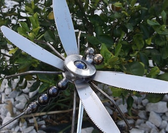 Pode incluir: Uma escultura de libélula de metal feita com talheres reciclados. A libélula tem uma gema azul no centro e é decorada com contas pretas. A escultura está sobre uma cama de cascalho branco.