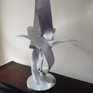 Könnte beinhalten: Eine silberne abstrakte Skulptur aus Metallblechen. Die Skulptur besteht aus mehreren gebogenen und angewinkelten Teilen, die miteinander verschweißt sind. Die Skulptur steht auf einem runden Sockel.
