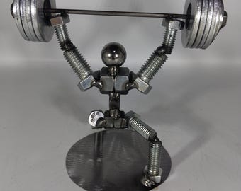 May include: A metal sculpture depicting a weightlifter. The figure is made from metal components like bolts, nuts, and washers. The weightlifter is lifting a barbell with circular weights. The sculpture is mounted on a circular base.