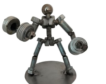 May include: A metal sculpture of a figure lifting weights, constructed from various steel hardware components including bolts, nuts, and washers. The figure features a spherical head, threaded limbs, and is mounted on a circular metal base.