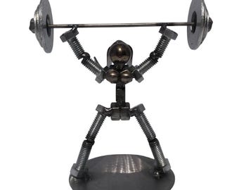 May include: A metal sculpture of a figure lifting a barbell, constructed from industrial hardware components including threaded bolts, nuts, and washers. The figure is mounted on a circular metal base, with a dark grey finish throughout.