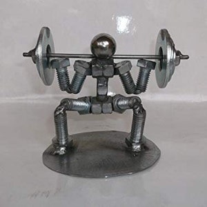 Könnte beinhalten: Eine Metallskulptur einer Person, die Gewichte hebt. Die Skulptur besteht aus Muttern, Schrauben und Unterlegscheiben. Die Person befindet sich in einer hockenden Position mit ausgestreckten Armen über dem Kopf. Die Gewichte bestehen aus Metallscheiben.