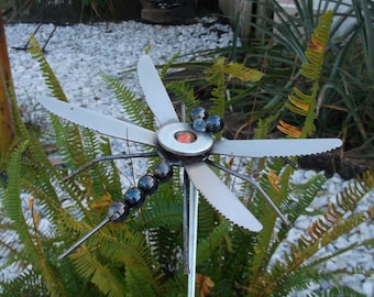 Pode incluir: Uma escultura de libélula de jardim de metal com um centro vermelho e contas pretas para os olhos. A escultura é feita de metal reciclado e tem um aspecto rústico.