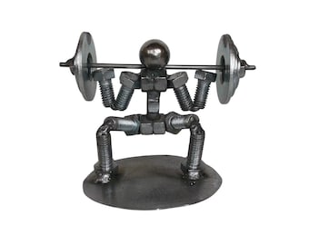 May include: A metal sculpture of a figure performing a squat while lifting a barbell. The figure is constructed from various steel hardware components, including bolts, nuts, and washers, with a spherical head and a flat circular base.