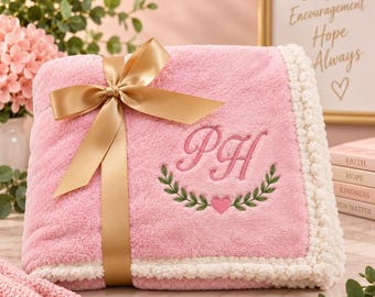 May include: A soft pink plush blanket featuring a cream-colored pom-pom border and a gold satin ribbon tied in a bow. The blanket is embroidered with the pink script letters PH above a green leaf wreath and a small pink heart.