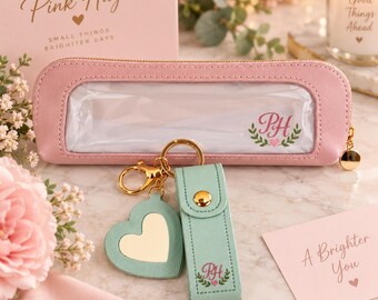 May include: A light pink pencil case with a clear window and a gold zipper, featuring a PH monogram. Attached is a gold keyring with a mint green heart-shaped mirror and a matching mint green leather-like strap with a gold snap and PH monogram.