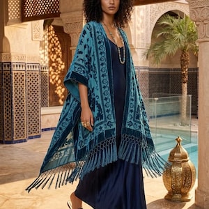 May include: A teal velvet kimono featuring a dark blue floral pattern, open front, and long fringe trim along the hem. The garment is worn over a solid dark blue sleeveless maxi dress. The kimono has wide sleeves and a structured border design.