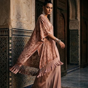 May include: A dusty rose velvet kimono featuring an intricate tonal floral pattern and long fringe trim along the hem, worn over a matching floor-length satin slip dress. The ensemble is accessorised with gold drop earrings and layered bracelets.