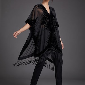 Puede incluir: Un kimono negro transparente con un patrón de terciopelo devoré, cuello en V, mangas cortas anchas y un dobladillo con flecos largos. La prenda se combina con pantalones negros y botines negros, creando un conjunto monocromático con textura.