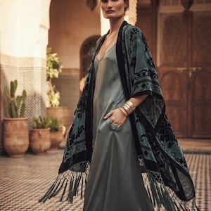 May include: A floor-length sage green slip dress with a V-neckline, paired with a sheer sage green kimono featuring black velvet floral patterns and long fringe trim along the hem. The ensemble includes gold-toned bangles and a turquoise ring.