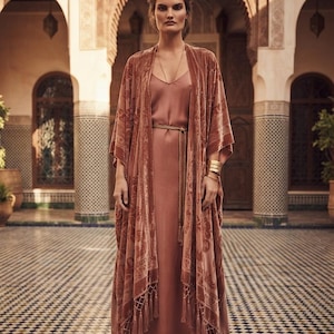 Könnte beinhalten: Ein bodenlanges Satin-Unterkleid in Altrosa, kombiniert mit einem passenden Samt-Kimono mit Ton-in-Ton-Blumenmuster, langen Fransen am Saum, einem dünnen Taillenband, einer schlichten Goldkette und gestapelten goldenen Armreifen am Handgelenk.