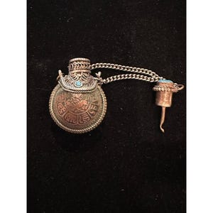 May include: A round metal snuff bottle featuring a copper-toned center with an embossed animal motif, surrounded by a silver-toned filigree border. A small turquoise stone is set at the top, and a matching stopper is attached by a silver chain.