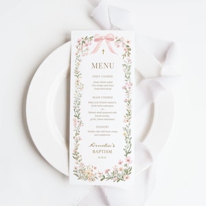 Può includere: Un biglietto del menu del battesimo con un design floreale. La carta presenta un fiocco rosa e una croce in alto, con le voci del menu elencate di seguito. Il menu comprende piatti come l'insalata di formaggio di capra, il carré di agnello e la torta alla vaniglia. La carta è su un piatto bianco con un nastro bianco.