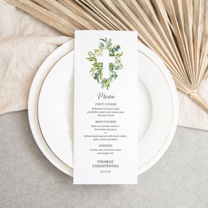 May include: A white menu card with a green floral cross design. The menu lists the courses for a christening: Ballantine of muscovy duck, baby rack of lamb, salmon steak, vanilla cake, and the date 01.10.21. The text "Thomas' Christening" is at the bottom of the card.