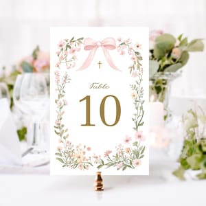 May include: A white table number card with a floral wreath design. The card features a pink bow, a gold cross, and the word "Table" above the number "10" in gold. The card is held by a gold metal holder.
