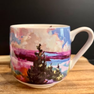 May include: A ceramic mug featuring a colorful painterly landscape design. The artwork depicts dark evergreen trees in the foreground against a vibrant sky filled with pink, purple, and blue clouds. The mug has a white interior and a white handle.