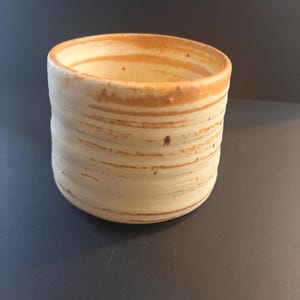May include: A cylindrical ceramic cup with a cream-colored base and horizontal, irregular brown stripes. The exterior surface features subtle, tactile ridges along the sides, and the rim is finished with a warm, earthy brown tone.