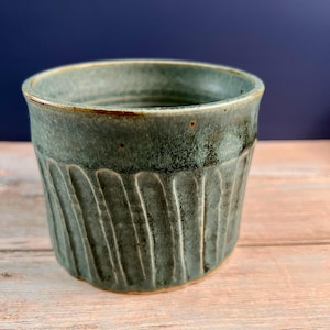 May include: A ceramic cup with a teal and green glaze featuring vertical fluted grooves along the exterior. The rim is smooth and rounded, while the base shows a hint of natural clay color. The surface has a mottled, textured finish.
