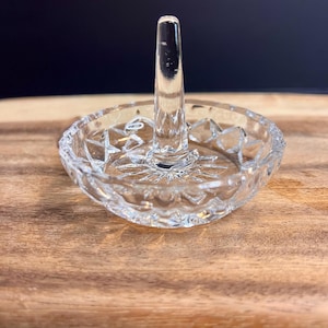 May include: A clear glass ring holder featuring a central vertical post for rings and a shallow, circular base with a faceted, starburst-patterned bottom and a scalloped, textured rim for holding small jewelry items securely.