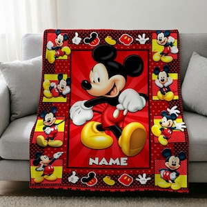 Könnte beinhalten: Eine rechteckige Fleecedecke mit einer großen zentralen Abbildung von Mickey Mouse auf einem roten Strahlenhintergrund und dem Wort NAME darunter. Der Rand enthält kleinere Mickey Mouse-Figuren in gelben Quadraten und roten Mustern.