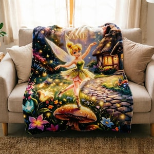 May include: A plush throw blanket featuring a colourful illustration of a blonde fairy with translucent wings standing on a large mushroom. The scene includes a stone path, a small cottage, and vibrant flowers. The word NAME is printed at the top.