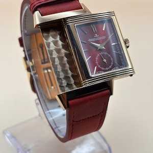 Op de afbeelding: Een rechthoekig goudkleurig polshorloge met een dieprode wijzerplaat met zonnestraalpatroon en de tekst Jaeger-LeCoultre. Het horloge heeft een bijpassende rode leren band en een draaibare kast met een gestructureerde, geschulpte metalen afwerking.