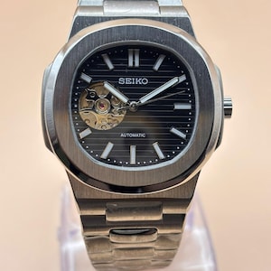 May include: A silver-toned stainless steel wristwatch with a black horizontal-lined dial, featuring the Seiko logo, an automatic movement, and an open-heart aperture at the nine o'clock position. The watch includes a matching metal link bracelet.