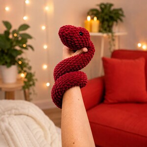 Puede incluir: Un juguete de serpiente de peluche rojo oscuro hecho de hilo grueso y suave. La serpiente presenta una cabeza redondeada con pequeños ojos oscuros incrustados y un cuerpo largo y enroscado que se envuelve alrededor del brazo. Textura gruesa.