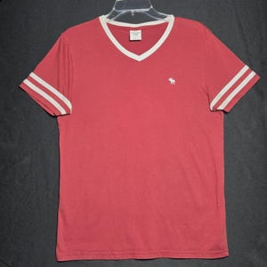 May include: A red V-neck t-shirt featuring white trim along the neckline and two white horizontal stripes on each short sleeve. A small white embroidered moose logo is positioned on the left chest area. The interior neck label reads Abercrombie.