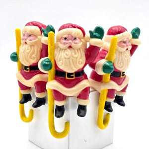 May include: Three identical Santa Claus figurines, each wearing a red suit with a black belt and white trim, green gloves, and black boots. Each Santa is holding a long, vertical yellow candy cane-shaped hook, designed to hang over an edge.