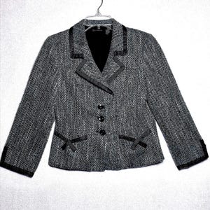 May include: A black and white tweed blazer featuring a notched lapel with black satin trim, three front buttons, and two decorative pockets with black satin ribbon accents. The long-sleeved jacket has a textured weave and a black interior lining.