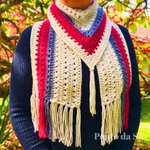 May include: A crochet scarf featuring a triangular neck section and long ends, crafted with alternating vertical stripes in cream, bright red, and muted blue. The scarf is finished with long, cream-coloured fringe tassels along the bottom edges.
