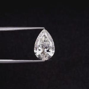May include: A pear-shaped, colorless, faceted gemstone with a brilliant cut, held by a pair of silver-toned metal tweezers. The stone displays multiple triangular and kite-shaped facets that reflect light, creating a clear, sparkling appearance.