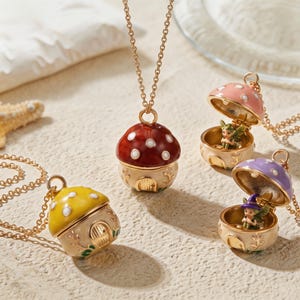 Puede incluir: Una colección de cuatro collares con colgante en forma de seta y cadenas doradas. Los colgantes tienen sombreros coloridos con puntos blancos y bases color crema con puertas arqueadas. Dos colgantes están abiertos, revelando pequeñas hadas.