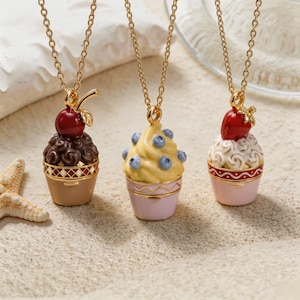 May include: Three gold-tone chain necklaces featuring pendant charms shaped like miniature desserts. The left pendant is a chocolate swirl with a cherry, the centre is yellow soft-serve with blueberries, and the right is white frosting with a strawberry.