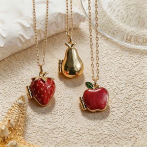 Peut inclure: Trois colliers en métal doré avec des pendentifs en forme de fruits. Un pendentif est une fraise rouge avec des graines blanches, un est une poire dorée lisse et un est une pomme rouge avec une feuille verte. Chaque pendentif est sur une chaîne dorée.