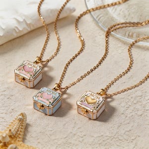 May include: Three gold-tone chain necklaces featuring square locket pendants. Each locket is white with floral accents and the word MOM printed on the front. The centre of each locket displays a heart in either pink or gold, suspended by a gold chain.