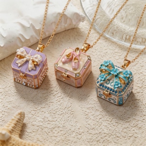 Può includere: Tre collane con pendenti quadrati su catenine dorate. Il pendente a sinistra è color lavanda con un fiocco decorato da perle. Quello centrale è color crema con scarpette da danza rosa. Quello a destra è bianco e blu con un fiocco floreale e cristalli blu.