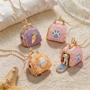 May include: Four gold-tone chain necklaces featuring small, hinged, handbag-shaped pendants with enamel finishes. One purple pendant has a carrot, one pink has a blue paw print, one orange has a cat, and one pink opens to reveal a small white dog figure.