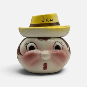 May include: A ceramic jam jar shaped like a head with a surprised expression, featuring rosy cheeks, brown hair, and a yellow hat lid with a brown band. The word Jam is written in black script on the front of the yellow hat. The jar is glossy.