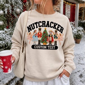 Könnte beinhalten: Ein beigefarbenes Sweatshirt mit Rundhalsausschnitt und einem Grafikdruck mit dem Text NUTCRACKER und CUSTOM TEXT. Das Design zeigt Illustrationen von zwei Ballerinas, einem Nussknacker, einem geschmückten Weihnachtsbaum, einem Mäusekönig und einem Soldaten.